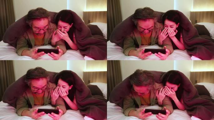 幸福亚洲甜蜜夫妇丈夫妻子享受智能手机电视节目节目一起在白色软床和毯子家庭室内背景