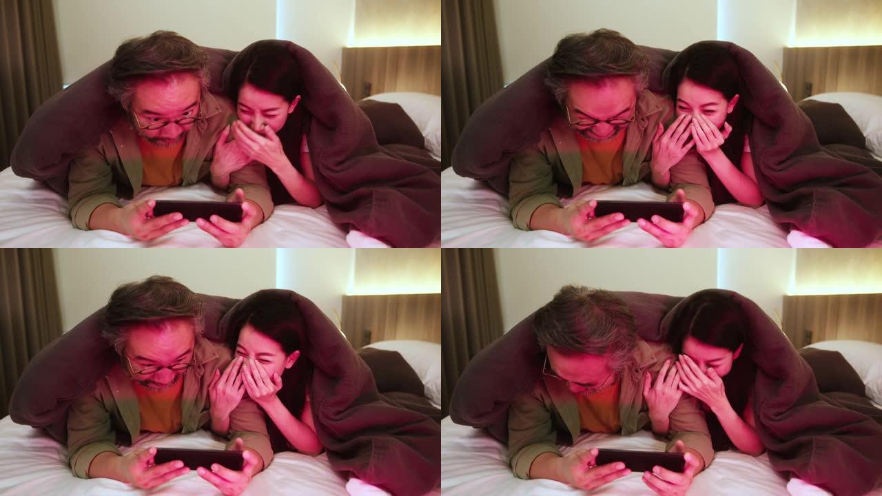 幸福亚洲甜蜜夫妇丈夫妻子享受智能手机电视节目节目一起在白色软床和毯子家庭室内背景