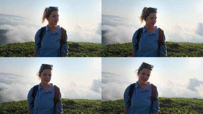 十几岁的女孩徒步旅行者站在山顶，站在云层上方，看着相机