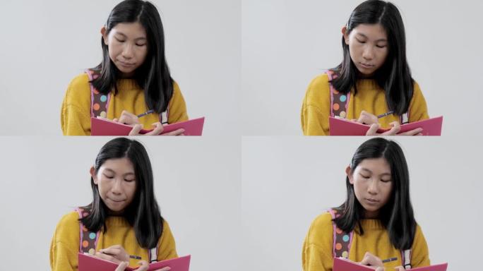 亚洲背包女学生穿着黄色毛衣写在粉红色的书上，在灰色的背景下，生活方式的概念。
