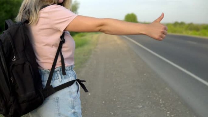 一个背着背包的女孩站在路边，竖起大拇指接车。搭便车。用手停车