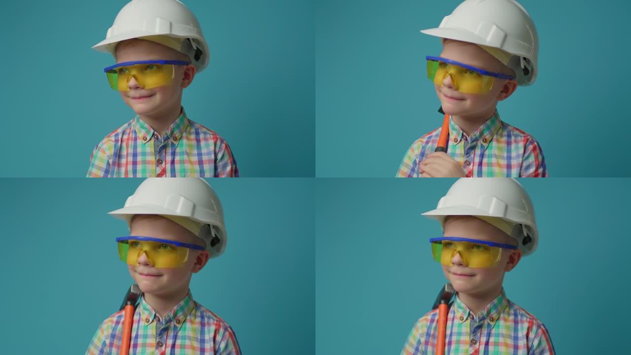 戴着白色安全帽和黄色防护眼镜的微笑男孩在蓝色背景上手持悍马。年轻的儿童建筑工程师。