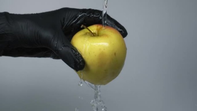 用水花在灰色背景上洗净手中的青苹果，慢动作。男人戴着黑色手套握住青苹果，在水流下洗净