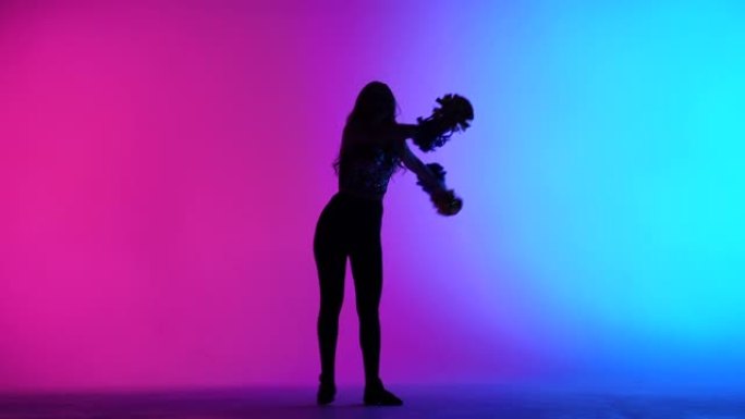苗条的啦啦队长的剪影，在工作室里，在粉红色的蓝色灯光的背景下，手中的绒球充满活力地跳舞。体育比赛中的