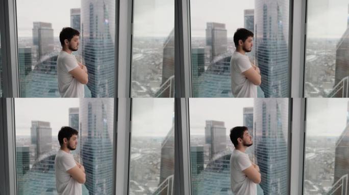 年轻人在顶层俯瞰城市的现代公寓里思考并看着窗外