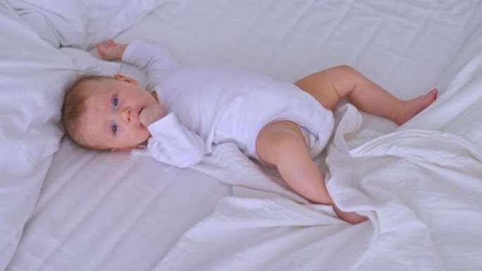 穿着白色紧身衣的新生婴儿吮吸嘴里的手指，躺在明亮房间的床上。可爱的蓝眼睛宝宝，童年。摄像机的俯视图。