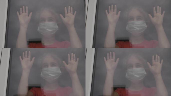 戴着防护面具的小女孩，悲伤地透过窗户看着担心新型冠状病毒肺炎封锁。戴着保护面具的孩子看着窗外的家。检