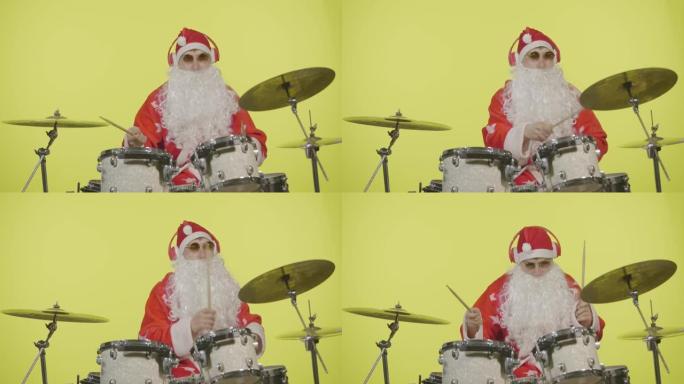 圣诞老人穿着节日服装，戴着胡须和太阳镜，在黄色工作室背景下打鼓。穿着狂欢节服装的人刘海鼓和打击钹创造