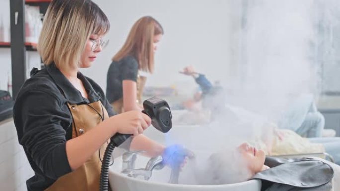 亚洲专业造型师或美发师使用纳米蒸汽枪头发处理来滋润客户的干燥头发。理发师在理发师或美容院为年轻女孩的