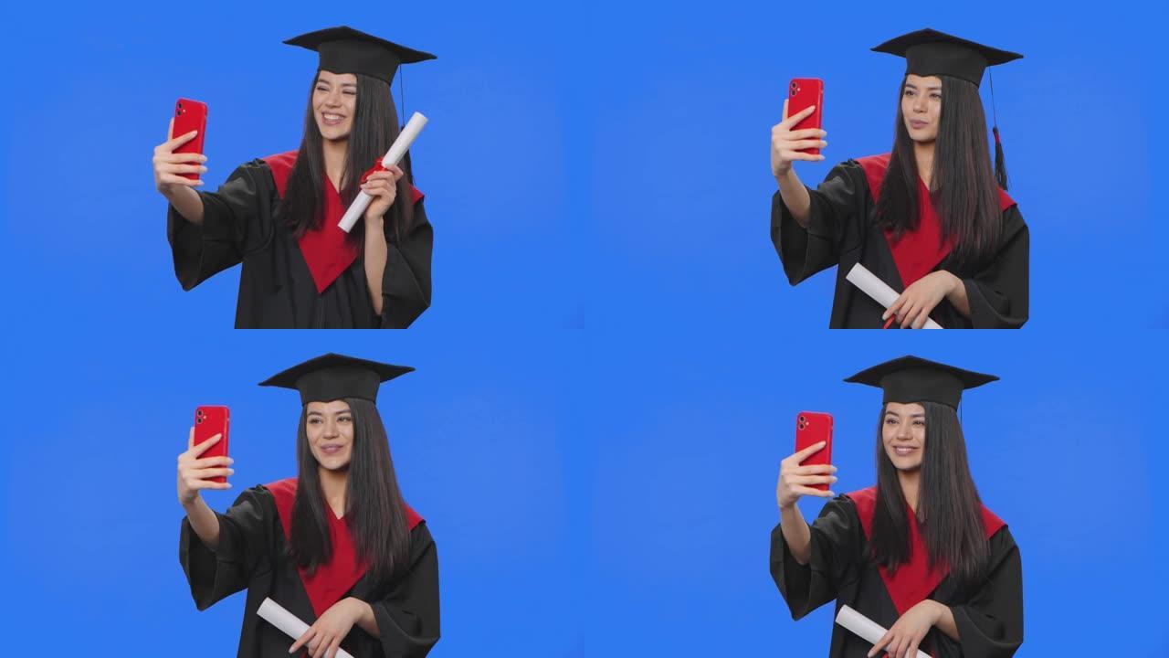 穿着毕业服装的女学生肖像，拿着文凭，用电话视频通话。年轻女子在工作室摆姿势，蓝屏背景。特写。慢动作准
