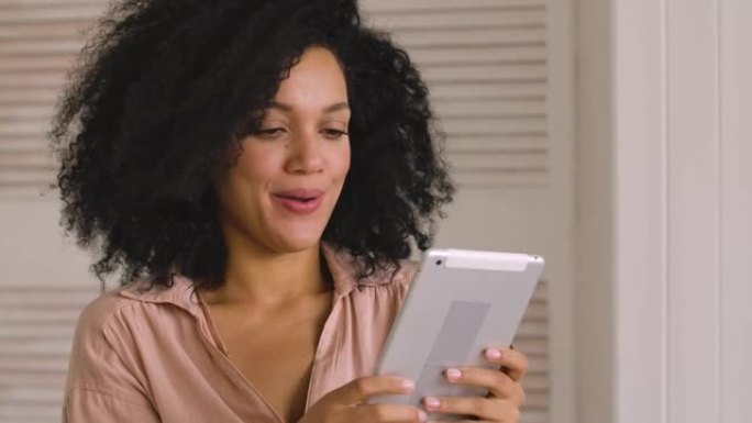 可爱的非洲裔美国妇女在数字平板电脑上寻找信息，并快乐地庆祝胜利，收到了好消息。黑人女性坐在灯光室。特