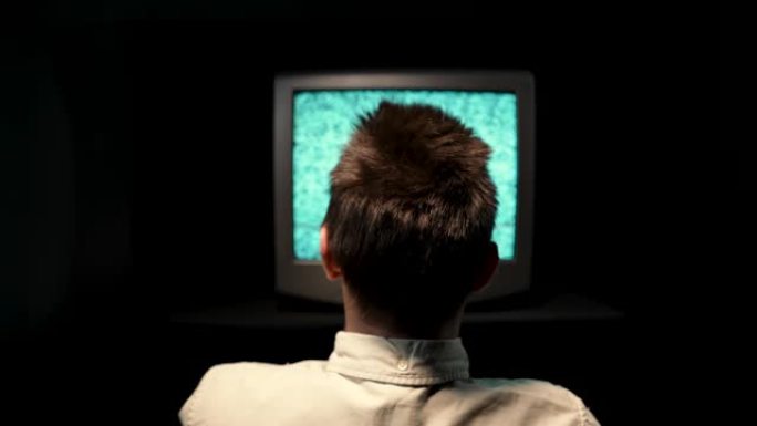 一个男人坐在一台旧电视前的后视图，在黑色背景的黑暗演播室中的电视监视器上受到噪音干扰。那个人在破电视