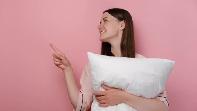 年轻漂亮的女人穿着睡衣，手指头指向一边，拿着白色枕头，在粉红色的工作室背景下孤立地摆姿势，为您的文本