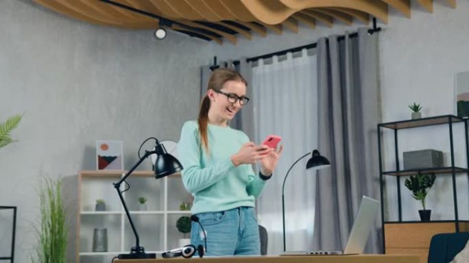 可爱的微笑快乐20岁的浅色头发女孩穿着便服站在客厅的桌子旁边，用她的手机看有趣的视频