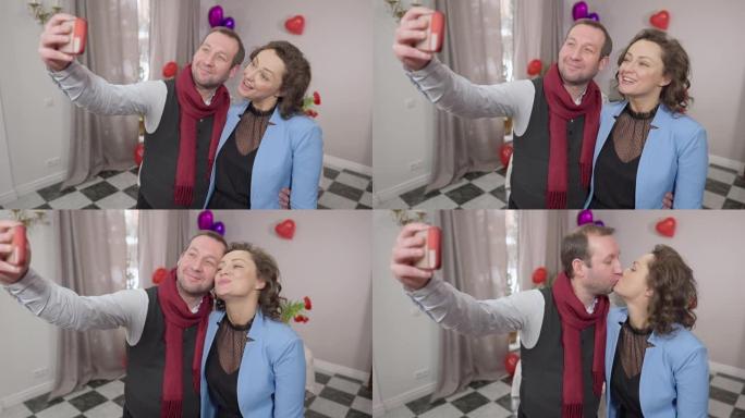 积极有趣的成年夫妇在情人节在室内自拍。微笑的爱的成年男人和女人在智能手机相机上做鬼脸。有趣和快乐的概
