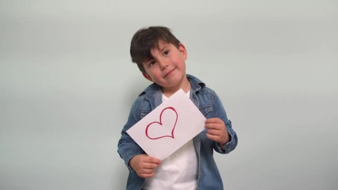 一个男孩拿着一张纸，画了一颗心，笑了一个开朗的孩子在灰蓝色的背景上显示一个带有心脏的标志开朗的孩子幸