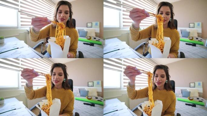 一个有趣的迷人年轻女子在电脑旁工作时在房间里吃意大利面。一个饥饿的年轻女子吃着美味的意大利面。