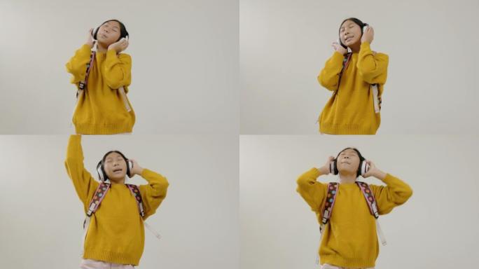 快乐的亚洲女学生穿着黄色毛衣跳舞唱歌，同时在灰色背景上使用无线耳机，生活方式理念。