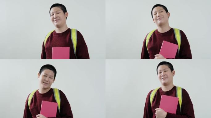 快乐的亚洲男生穿着红色毛衣，拿着粉红色的书，看着灰色背景上的相机，生活方式的概念。