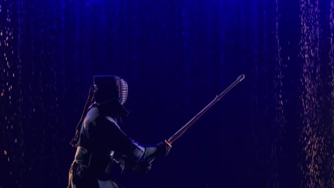日本剑道战士的侧视图，带着竹剑和传统服装，正在雨滴和蓝光的黑暗工作室中训练。特写。慢动作