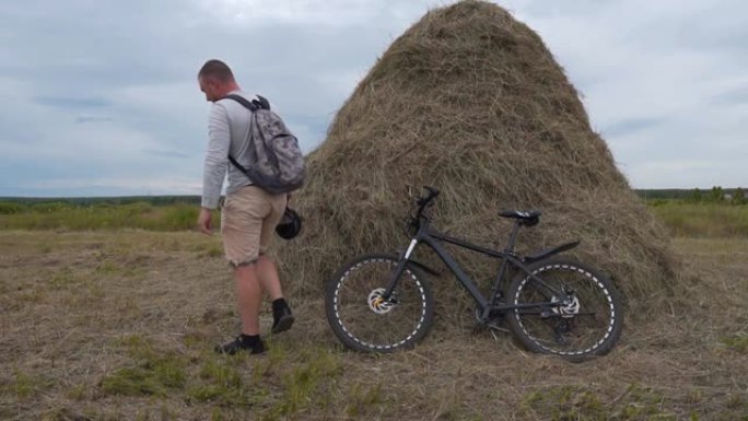 一个骑自行车的人在田野的干草堆附近停下来休息一下。他从自行车上爬下来，将其靠在干草堆上，然后脱下背包