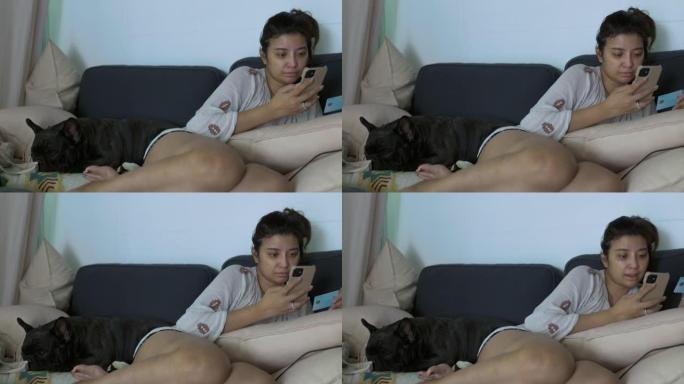 亚洲女人使用智能手机和可爱的可爱的黑色法国斗牛犬喜欢在家睡在沙发上。