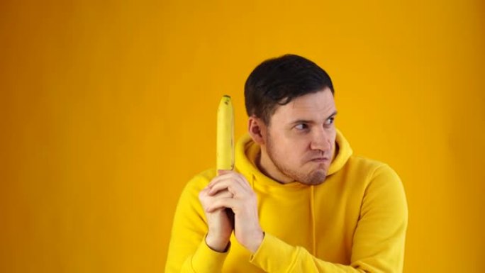 黄色背景上有香蕉的年轻人的肖像。穿着黄色连帽衫的帅哥的特写镜头玩水果，把它想象成武器