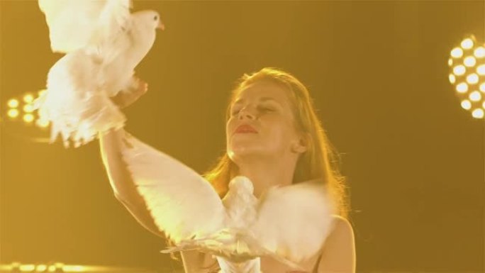 女教练与受过训练的白色鸽子一起表演，鸽子坐在手上并拍打翅膀。在明亮的聚光灯背景下，在黑暗的工作室里与