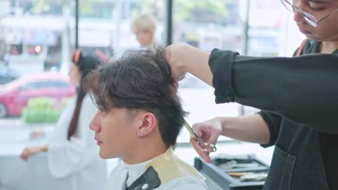 亚洲专业男性发型师在沙龙中梳理和使用剪刀剪掉年轻顾客的头发。坐在商店里看着镜子的男人微笑着与专家发型