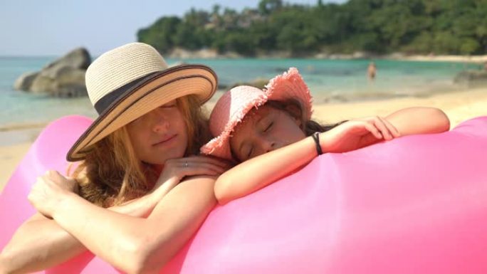 戴着草帽的妈妈和女儿在躺在粉红色充气沙发上的海滩上享受闲散