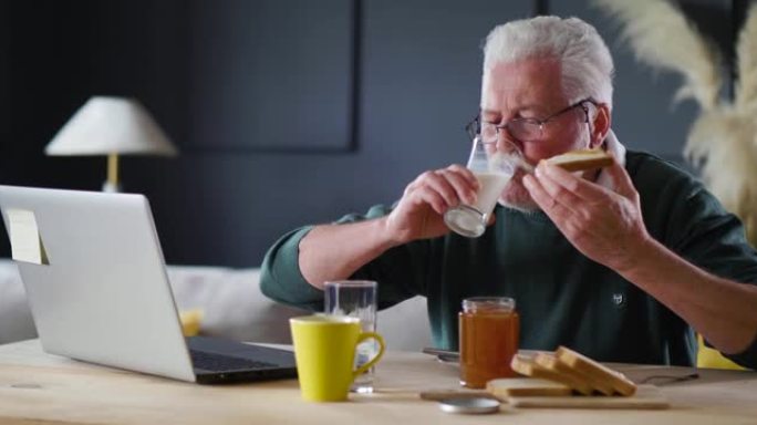 老人吃早餐，看着他的笔记本电脑。留着胡子的爷爷早餐吃果酱喝牛奶