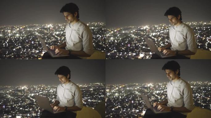 一名年轻的印度男性坐在顶部，晚上用笔记本电脑对着照明的城市景观