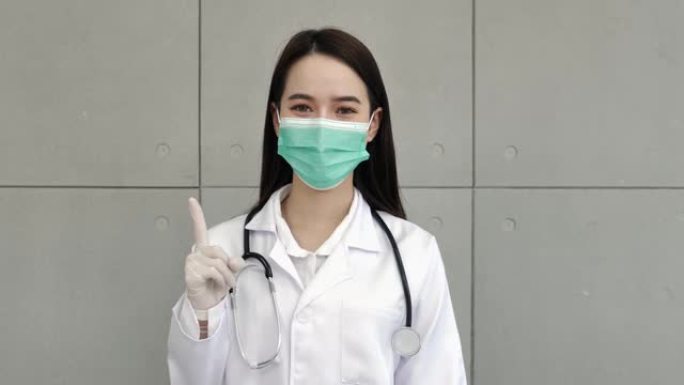 一名亚洲女医生或护士穿着制服，配有听诊器，戴着医用口罩，戴着手套，站着举起右手和食指。