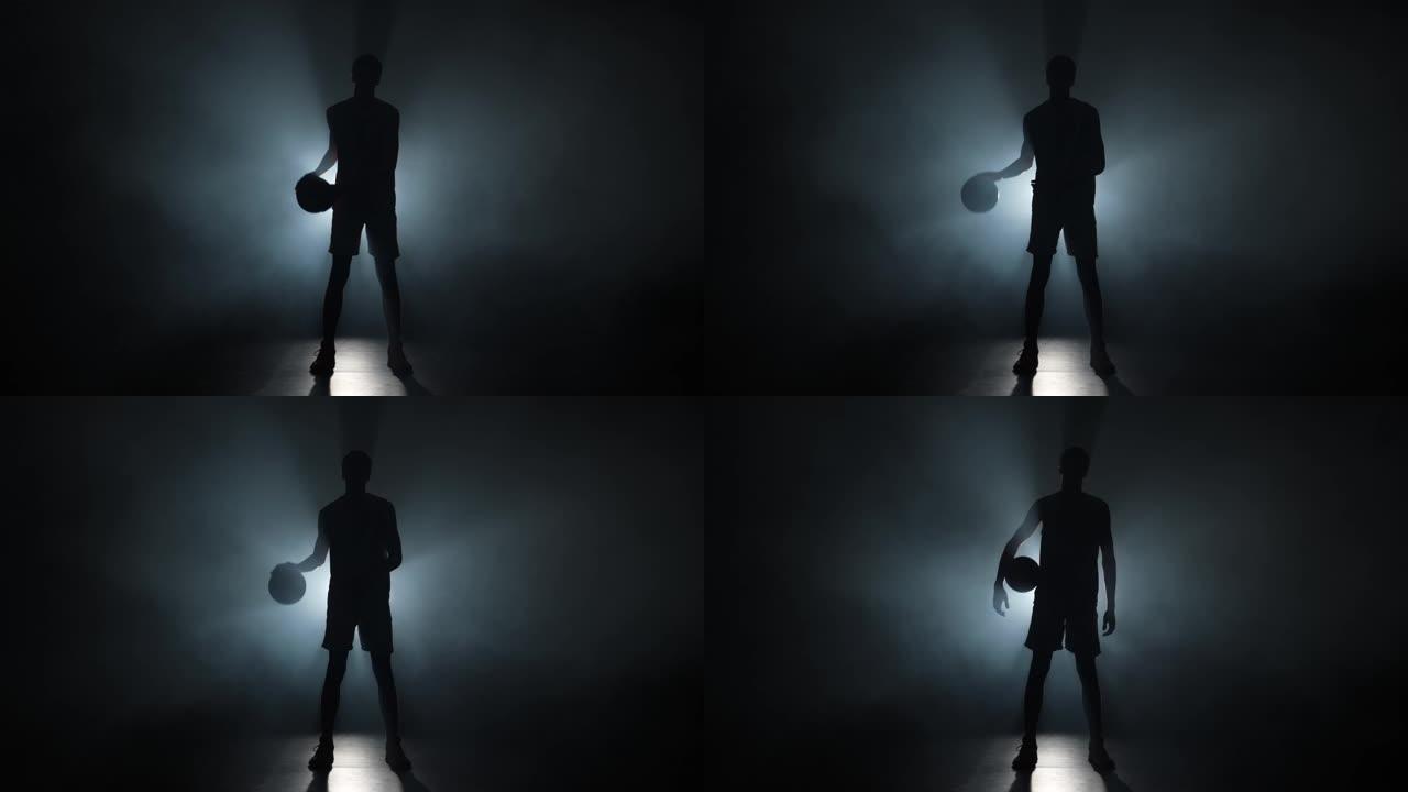 一个年轻人在黑暗的演播室背景上带烟和背光运球篮球的剪影。穿着运动服的高个子年轻运动员在地板上击球。慢