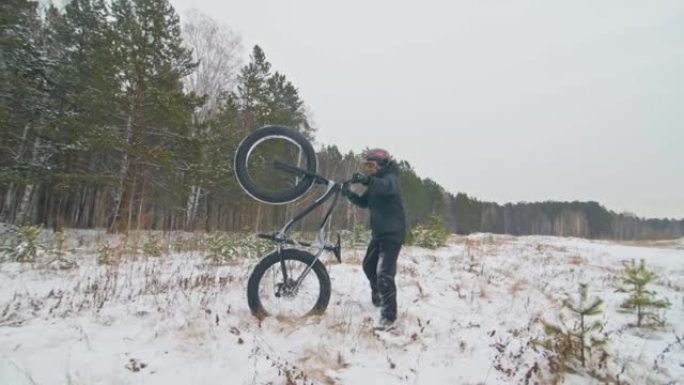 专业极限运动员骑自行车的人在户外举起一辆胖自行车。骑自行车的人留在冬天的雪林里。男子带着戴着头盔和眼