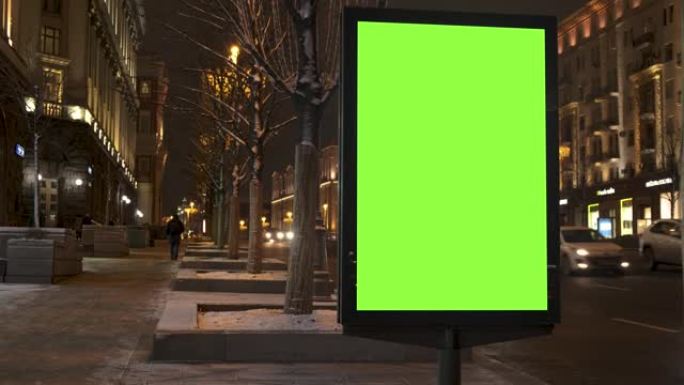 一条绿屏广告牌位于人们行走的人行道上，紧挨着一条繁忙的街道。汽车在晚上行驶。