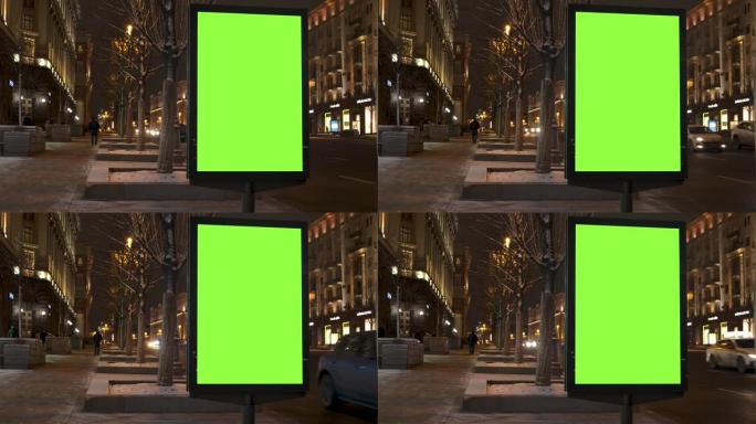 一条绿屏广告牌位于人们行走的人行道上，紧挨着一条繁忙的街道。汽车在晚上行驶。