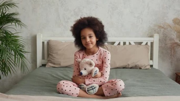 相机放大脸小非洲裔美国女孩拥抱她最喜欢的泰迪熊。穿着睡衣的少女坐在卧室的床上，室内明亮。慢动作。特写