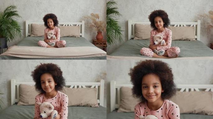 相机放大脸小非洲裔美国女孩拥抱她最喜欢的泰迪熊。穿着睡衣的少女坐在卧室的床上，室内明亮。慢动作。特写
