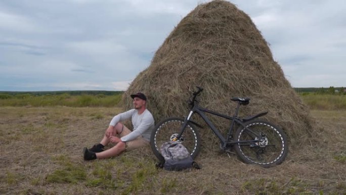 骑自行车的人坐在干草堆旁，享受周围的自然风光，从侧面看。骑自行车的健康人。自由旅行者骑自行车旅行