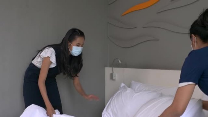 两名戴着医用口罩的年轻女仆在酒店房间的床上换床单