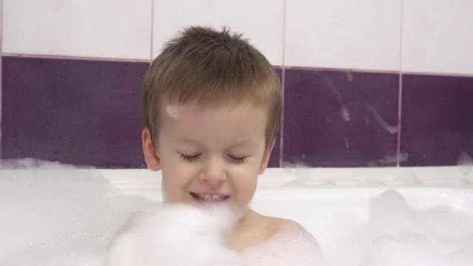 一个快乐微笑的小男孩的特写镜头，浴缸里有很多泡沫。他拍手泡沫。