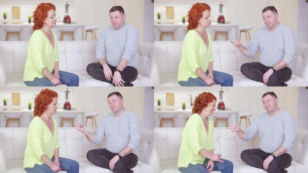 红发高加索女人和黑发男人用小手指做和平。正面的粉红色发誓在家里的房子里争论夫妻。坐在沙发上微笑的情侣