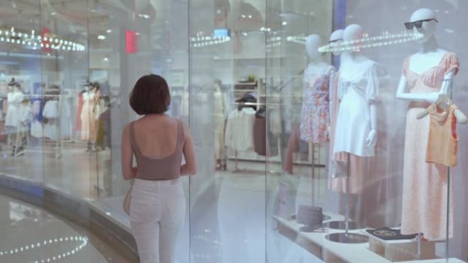 时尚女人步行经过商场的现代玻璃纺织服装店。百货商店的顾客步行通行证橱窗展示和人体模型娃娃。