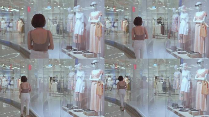 时尚女人步行经过商场的现代玻璃纺织服装店。百货商店的顾客步行通行证橱窗展示和人体模型娃娃。
