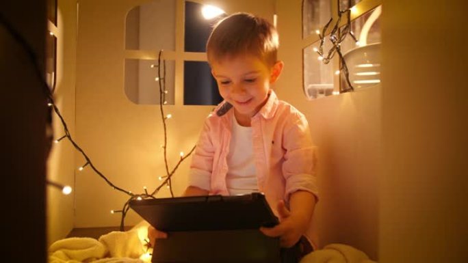 快乐微笑的男孩的肖像使用平板电脑，并在晚上在他的玩具纸板屋或帐篷里玩耍时浏览互联网。儿童教育和夜间学