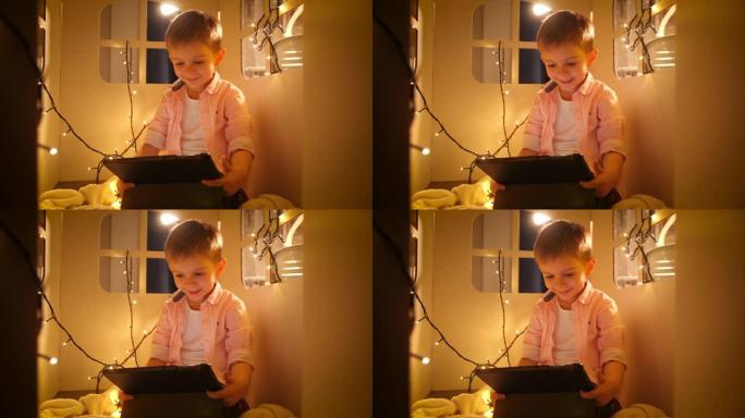 快乐微笑的男孩的肖像使用平板电脑，并在晚上在他的玩具纸板屋或帐篷里玩耍时浏览互联网。儿童教育和夜间学