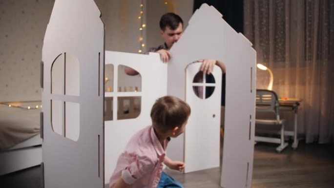 快乐微笑的小男孩在他的房间里玩耍，而父亲则在组装玩具纸板屋。家人在一起玩得开心。孩子们帮助父母。