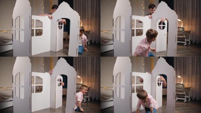快乐微笑的小男孩在他的房间里玩耍，而父亲则在组装玩具纸板屋。家人在一起玩得开心。孩子们帮助父母。