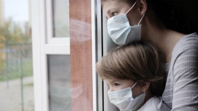 成熟的女人带着她的儿子，戴着防护面具，悲伤地透过窗户看着担心新型冠状病毒肺炎封锁。戴着保护面具的家庭